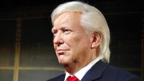 Portrait of an older man with white hair in a dark suit and red tie, wearing an American flag lapel pin.