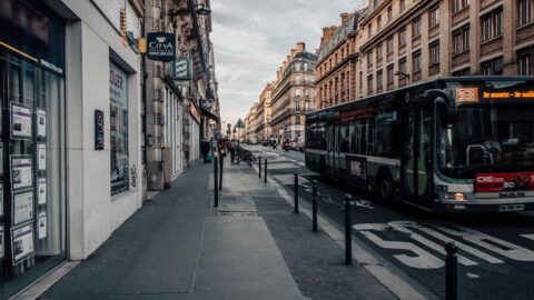 Urban European street scene with a city bus on the right, storefronts along the left, and tall brick buildings under a gray, overcast sky.