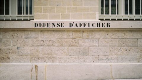 Stone wall with a plaster ledge bearing the French sign 'DÉFENSE D'AFFICHER', indicating posting is prohibited, with barred windows above.