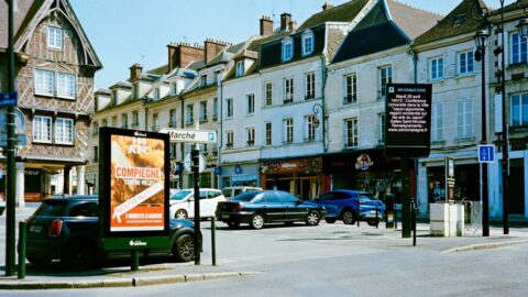 European town square with historic buildings, parked cars, and two large advertising boards along a pedestrian area (one board shows a poster for a Compiegne event).