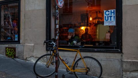 Yellow bicycle locked to a pole in front of a cafe window with warm interior lights and people inside, on a gray urban street.
