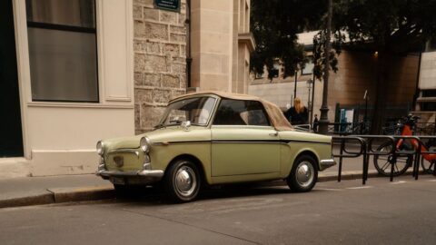 Vintage light-green convertible with tan top parked on a city street beside a stone building.