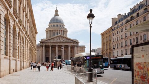 Street view in Paris toward the Panthéon with a domed capitol building, pedestrians, and tour buses along the curb.