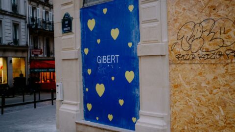 Blue storefront panel with yellow hearts and the word 'GIBERT' on a Paris street, adjacent to graffiti-covered plywood wall.