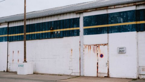 White brick warehouse with a rusted metal door, blue stripe, and a no-parking sign on the wall railings.