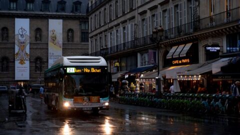 Nighttime Paris street; a bus with headlights on drives along a wet road past outdoor cafés and a line of green bikes outside buildings.