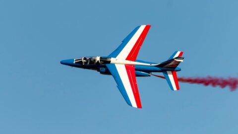 Blue aerobatic jet with red, white, and blue stripes flying left-to-right against a clear sky, trailing red smoke.