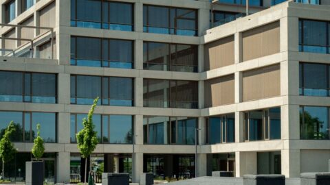 Modern multi-story office building with a beige concrete facade and large blue-tinted windows, with small trees and a paved plaza in the foreground.