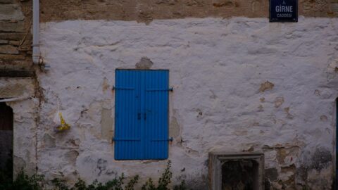 White stucco wall with a bright blue shuttered window, a street sign reading Girne Caddesi, and a concrete opening at ground level; a yellow bag hangs from a pipe on the left.