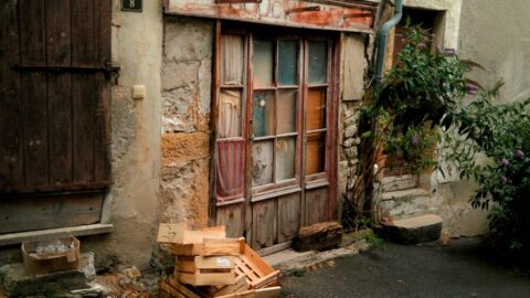 Rustic doorway with chipped wood and multicolored glass panes, stone wall, and a wooden crate piled on the ground outside.
