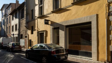 Sunlit street in a European town with a yellow building and a large storefront window; a black car parked at the curb in front of the shop.